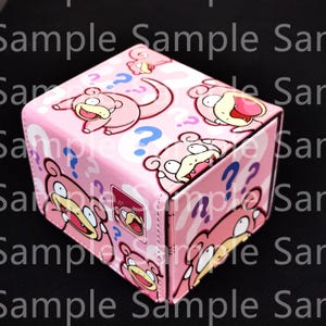 May include: A pink, cube-shaped box featuring cartoon illustrations of a Slowpoke character and question marks. The box has a repeating pattern of the character and question marks. The box is a light pink color.