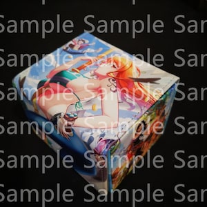 May include: A cube-shaped storage box featuring a colorful anime illustration. The artwork depicts a woman in a bikini on a beach scene. The box has a blue and white color scheme with additional character art on the sides.