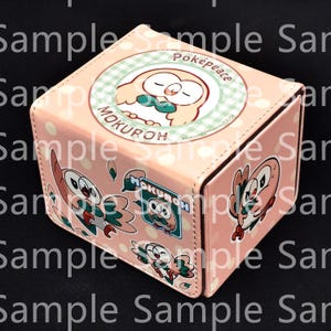 May include: A light pink rectangular storage box featuring a repeating white polka dot pattern and multiple illustrations of the character Mokuroh. The top lid displays a circular graphic with the text Poképeace and MOKUROH, while the side shows stickers.