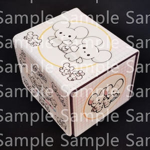 May include: A rectangular storage box with a white grid pattern and illustrations of small white mice. The top features two mice inside a yellow circle, while the side displays a group of mice in a similar circle. The box has a light color palette.