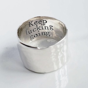 May include: A wide silver ring with a hammered exterior finish. The interior surface is engraved with the black text "Keep fucking going" and the small maker's mark "TB&Co". The band features an open, overlapping design at the front.