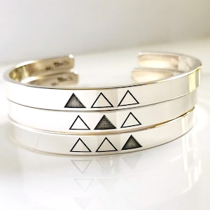 May include: Three silver bangle bracelets with a repeating pattern of black triangles.