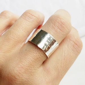 May include: A wide silver band ring featuring a hammered texture on the outer surface. The metal has a polished, reflective finish that catches the light, creating subtle variations in brightness across the curved surface of the band.