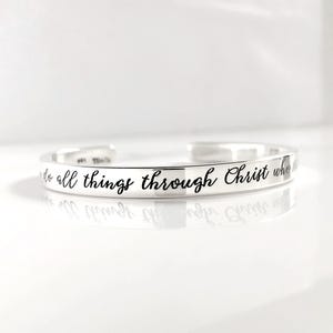 May include: A silver-toned metal cuff bracelet featuring a polished finish and an open-ended design. The exterior surface is engraved with the black script text, "I can do all things through Christ who". The bracelet has a reflective, smooth surface.