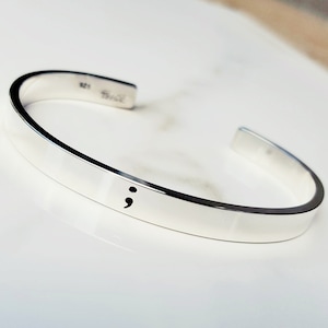Könnte beinhalten: Ein silbernes Armreif-Armband mit einem Semikolon, das auf der Vorderseite graviert ist. Das Armband ist schlicht gehalten und eine großartige Möglichkeit, die Sensibilisierung für psychische Gesundheit zu unterstützen.