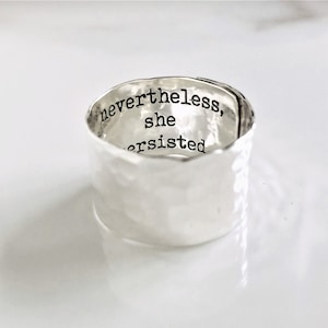 Könnte beinhalten: Ein silberner Ring mit gehämmerter Textur. Die Innenseite des Rings ist mit dem Text "nevertheless, she persisted." graviert.