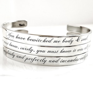 May include: A set of three silver-toned metal cuff bracelets stacked together. Each bracelet features engraved black cursive text. The inscriptions read: You have bewitched me body & soul, you must know, surely, you must know it was all for you, and completely and perfectly and incandescently happy.