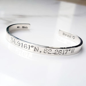 May include: Silver cuff bracelet with the coordinates 34.9161°N, 82.2617°W engraved on it. The bracelet also has the word "Home" and a heart symbol engraved on it.