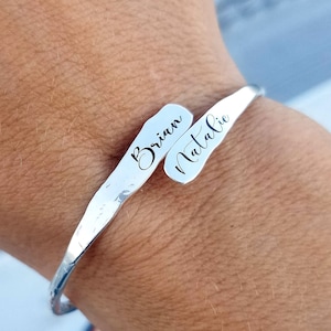 May include: A silver-colored cuff bracelet with the names "Brian" and "Natalie" engraved in a cursive font. The bracelet has a hammered texture and a curved, open design.