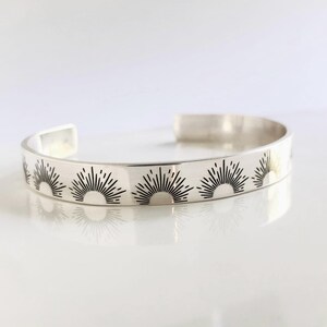 May include: A silver cuff bracelet with a repeating sunburst pattern in black. The open-ended bracelet is approximately 0.5 inches wide. The sunburst design is evenly spaced around the bracelet.