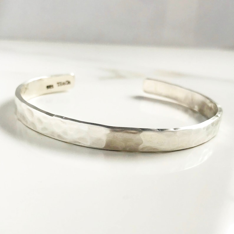 Puede incluir: Un brazalete de plata martillado. El brazalete es liso y tiene una forma ligeramente redondeada.