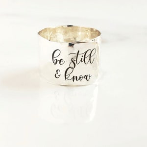 May include: Silver ring with a hammered finish and the inscription "be still & know" in black lettering.