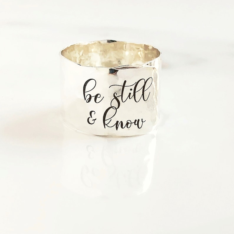 May include: Silver ring with a hammered finish and the inscription "be still & know" in black lettering.