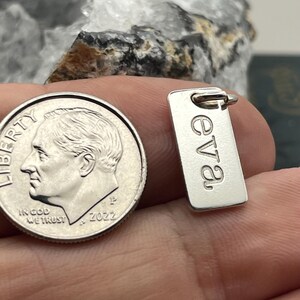 May include: A small rectangular silver-toned metal charm featuring the engraved lowercase text "eva" oriented vertically, positioned next to a United States dime for scale, showing the charm's compact size relative to the coin.