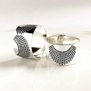 May include: Two silver rings with a black graphic design. The design is a stylized representation of a collar. One ring is upright, and the other is tilted on its side. The rings are on a white surface.