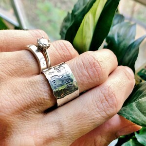 May include: A silver ring with the words "Be Still & Know" engraved on it. The ring is worn on a finger with a diamond ring on the finger next to it.