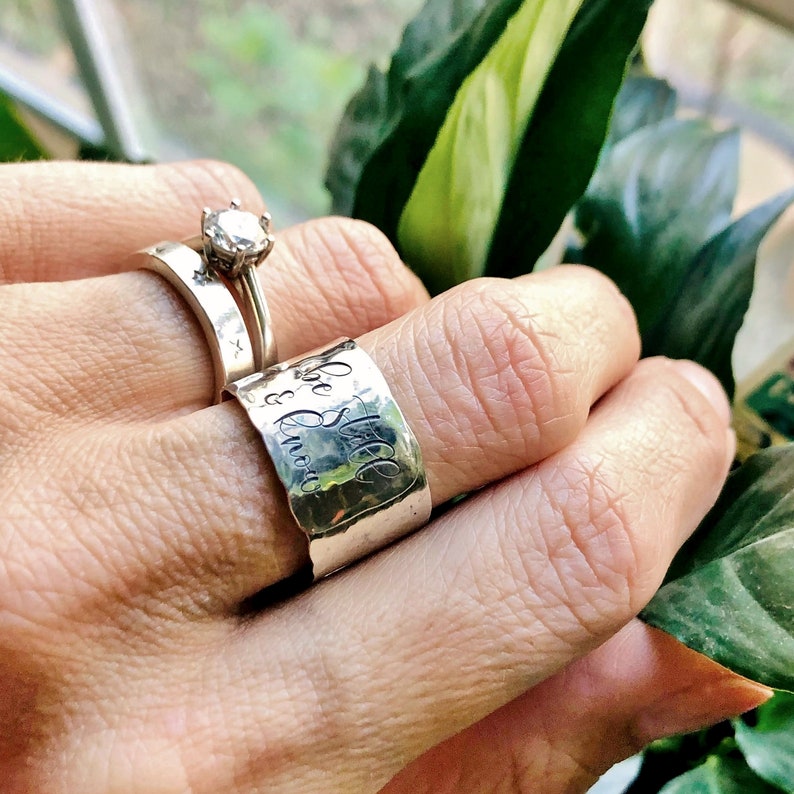 May include: A silver ring with the words "Be Still & Know" engraved on it. The ring is worn on a finger with a diamond ring on the finger next to it.
