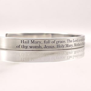 May include: A silver-colored cuff bracelet with the engraved text "Hail Mary, full of grace. The Lord is with of thy womb, Jesus. Holy Mary, Mother of God." The bracelet reflects the text below.