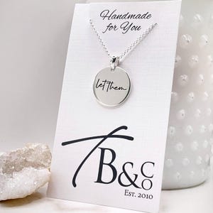Könnte beinhalten: Silberne Halskette mit einem runden Anhänger, auf dem "let them" steht. Die Halskette liegt auf einer weißen Karte mit den Worten "Handmade for You" und "B&C Est. 2010" darauf.