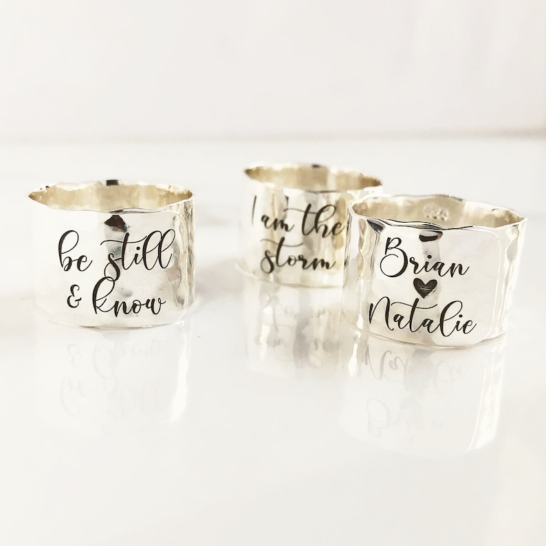 May include: Three silver rings with different engraved messages. The first ring says "be still & know". The second ring says "I am the storm". The third ring says "Brian Natalie" with a heart symbol between the names.
