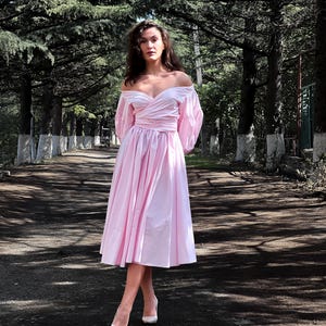 Op de afbeelding: Een lichtroze off-the-shoulder jurk met een getailleerde taille en een uitlopende rok. De jurk heeft lange, pofmouwen en wordt gedragen met witte hoge hakken. De sc&egrave;ne speelt zich buiten af, op een pad omzoomd door bomen.