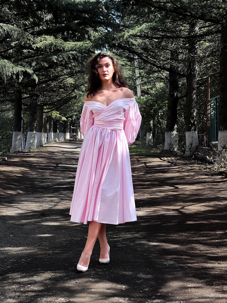 Op de afbeelding: Een lichtroze off-the-shoulder jurk met een getailleerde taille en een uitlopende rok. De jurk heeft lange, pofmouwen en wordt gedragen met witte hoge hakken. De sc&egrave;ne speelt zich buiten af, op een pad omzoomd door bomen.