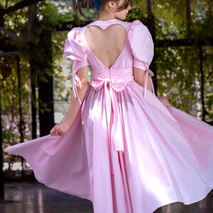 May include: A pink dress with a heart-shaped cutout in the back. The dress has a full skirt and is tied at the waist with a bow. The model is wearing black heels.