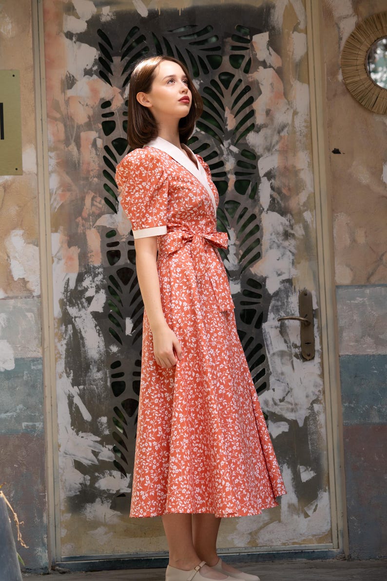 Pode incluir: Uma mulher vestindo um vestido coral com estampa floral branca. O vestido apresenta mangas curtas bufantes, gola branca e cinto combinando. O vestido, com cerca de 100 cm de comprimento, &eacute; usado com sapatos bege.