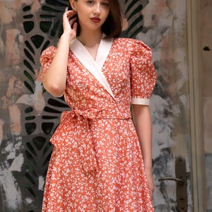 Pode incluir: Um vestido envelope coral com um padr&atilde;o floral branco. O vestido apresenta uma gola e punhos brancos, uma faixa de cintura a condizer e mangas curtas bufantes. O vestido &eacute; at&eacute; ao joelho.