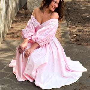 Op de afbeelding: Een lichtroze off-the-shoulder jurk met volumineuze mouwen en een volle rok. De jurk is gemaakt van een glanzende stof en heeft een getailleerd lijfje. Het model lacht en zit op een stenen ondergrond.
