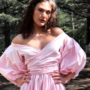 Op de afbeelding: Een lichtroze off-the-shoulder jurk met een wikkelbustier en een volle rok. De jurk heeft volumineuze, geplooide mouwen en is in de taille getailleerd. Het model staat buiten met een bosachtergrond.