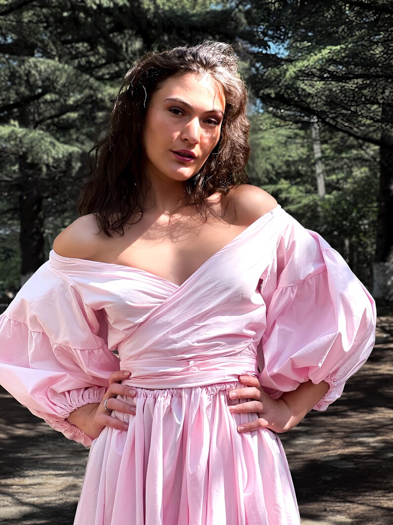 Op de afbeelding: Een lichtroze off-the-shoulder jurk met een wikkelbustier en een volle rok. De jurk heeft volumineuze, geplooide mouwen en is in de taille getailleerd. Het model staat buiten met een bosachtergrond.