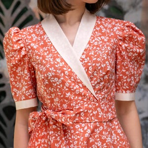 Pode incluir: Vestido envelope coral com estampa floral branca. O vestido tem gola creme, mangas curtas e cinto a condizer. O padr&atilde;o floral &eacute; composto por flores e folhas. Estilo vintage.