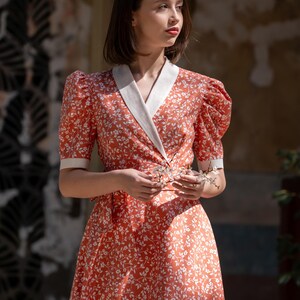 Pode incluir: Vestido coral com padr&atilde;o floral branco. O vestido apresenta mangas curtas bufantes, um corpete envolvente com gola branca e uma saia fluida. A modelo segura um pequeno ramo de flores brancas.