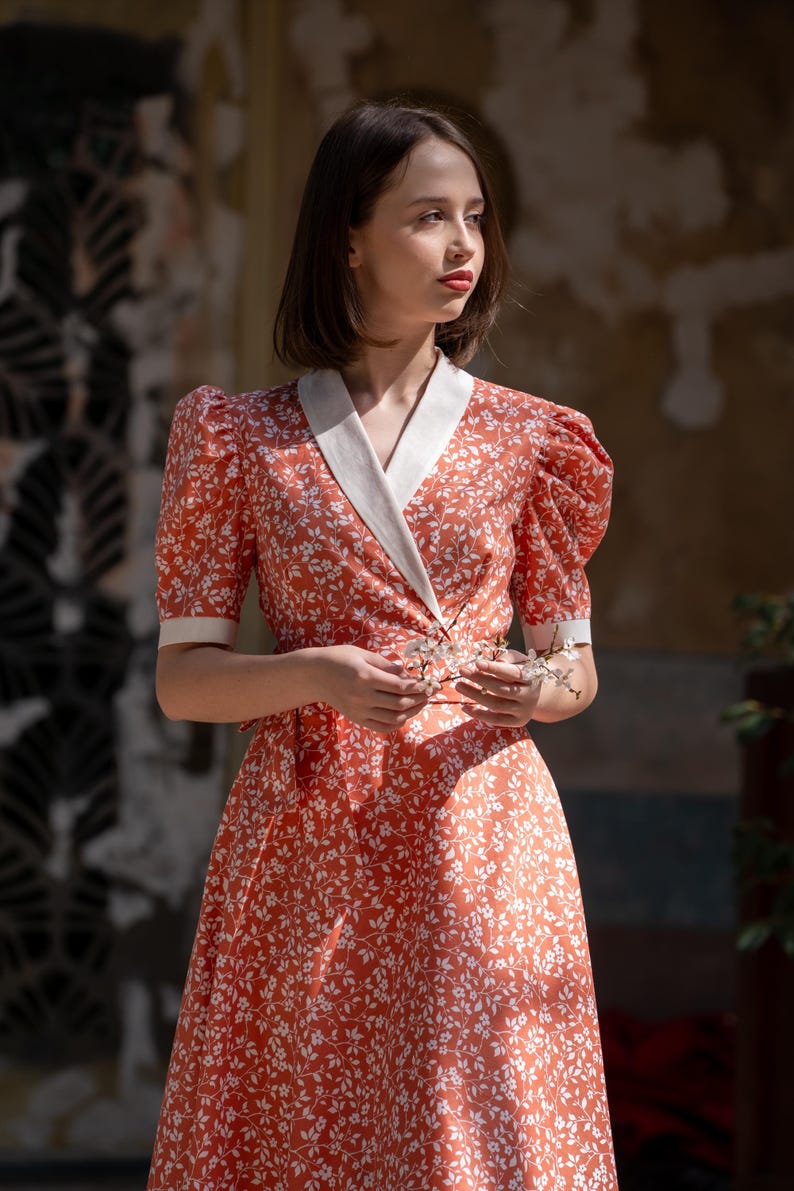 Pode incluir: Vestido coral com padr&atilde;o floral branco. O vestido apresenta mangas curtas bufantes, um corpete envolvente com gola branca e uma saia fluida. A modelo segura um pequeno ramo de flores brancas.