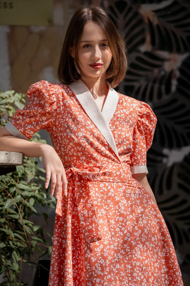 Pode incluir: Um vestido envelope em um tom laranja vibrante, adornado com um delicado padr&atilde;o floral branco. O vestido apresenta mangas curtas bufantes, uma gola e punhos brancos contrastantes e uma faixa combinando na cintura. O vestido &eacute; feito de tecido leve.
