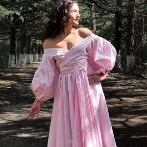 Op de afbeelding: Een lichtroze off-the-shoulder jurk met volumineuze mouwen en een getailleerde taille. De jurk heeft een zwierige rok die tot halverwege de kuit valt. Het model staat op een pad omringd door bomen.