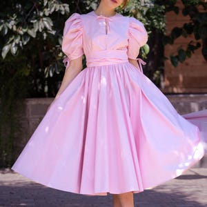 May include: A woman wearing a pink dress with puffed sleeves and a cinched waist. The dress has a full skirt and is made of a lightweight fabric. She is standing on a brick walkway.