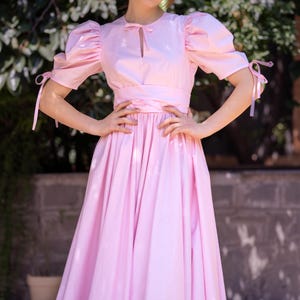 May include: A pink midi dress with puffed sleeves and a bow at the neckline. The dress has a fitted bodice and a full skirt.