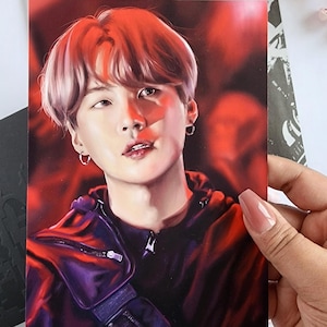 May include: A portrait print featuring a person with red hair and a black jacket. The print is held by a hand with long nails. The background is a blurred red. The text "Agust D" is visible on a paper behind the print.