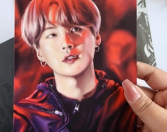May include: A portrait print featuring a person with red hair and a black jacket. The print is held by a hand with long nails. The background is a blurred red. The text "Agust D" is visible on a paper behind the print.