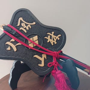 Puede incluir: Un objeto decorativo negro con caracteres y acentos japoneses dorados. Una cuerda roja está atada alrededor del objeto, con borlas rojas en el extremo. El objeto se exhibe sobre un soporte negro.