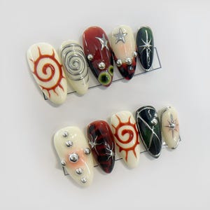 May include: Set of ten false nails with various designs. The nails are white with red, black, and green accents. Some nails feature a spiral design, others have a star, and some have a sun design.