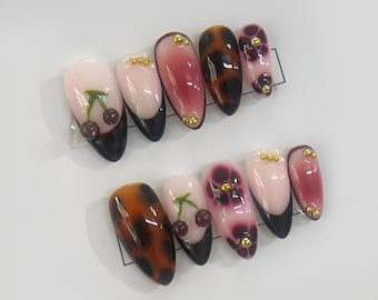 May include: Set of ten false nails with different designs. Some nails have a pink base with a black tip, others have a leopard print, and some have a floral design with a cherry.