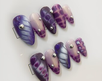 Uñas postizas abstractas con gel morado y blanco floreciente, con tachuelas plateadas, juego de uñas único y moderno.