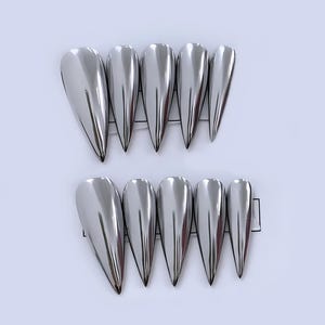 Puede incluir: Diez uñas postizas plateadas y metálicas con diseño puntiagudo. Dispuestas en dos filas de cinco, cada una con forma de lágrima y punta afilada. Tienen un acabado reflectante, similar a un espejo.