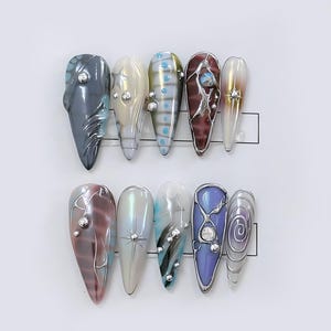 May include: A set of ten long, almond-shaped artificial nails. Each nail features a unique design with various colors, including gray, white, blue, and purple, and embellished with silver accents and rhinestones. The nails are arranged on a white surface.
