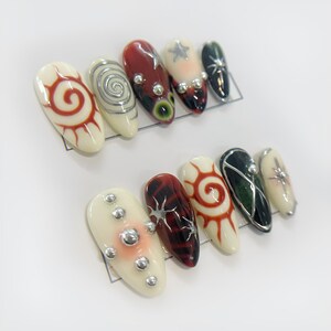 May include: Set of ten false nails with various designs. The nails are white with red, black, and silver accents. Some nails feature a spiral design, while others have a sun or star design.