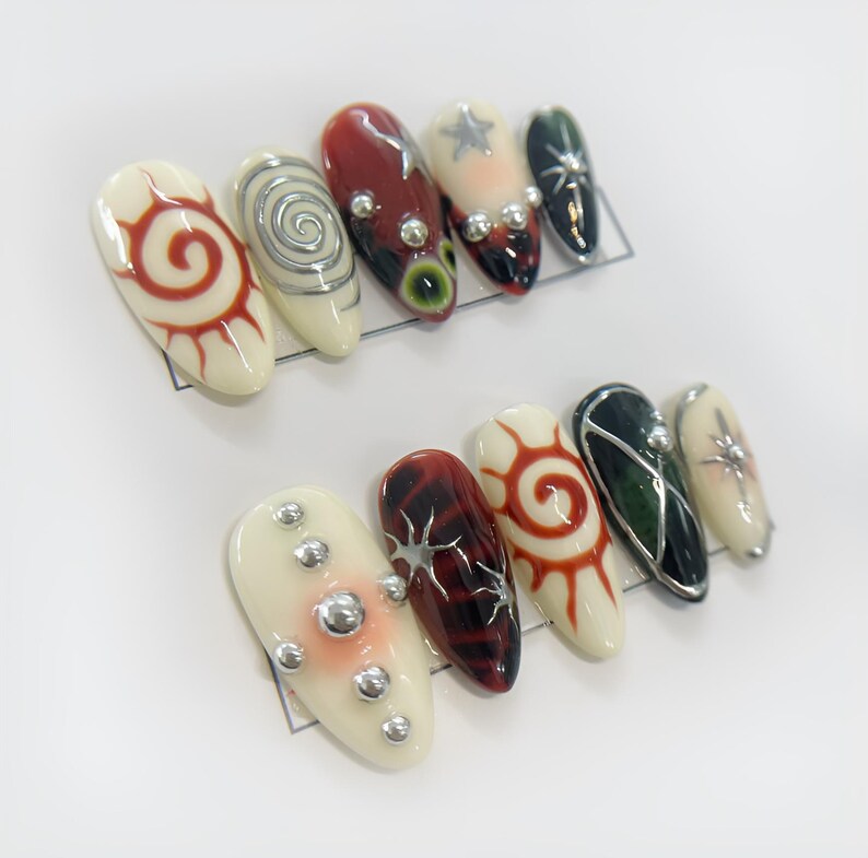 May include: Set of ten false nails with various designs. The nails are white with red, black, and silver accents. Some nails feature a spiral design, while others have a sun or star design.
