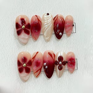 以下が含まれることがあります： 赤と白のグラデーションデザインの付け爪10本セット。一部の爪には金色のセンターを持つ赤い花が描かれており、他の爪には白い渦巻き模様が描かれています。